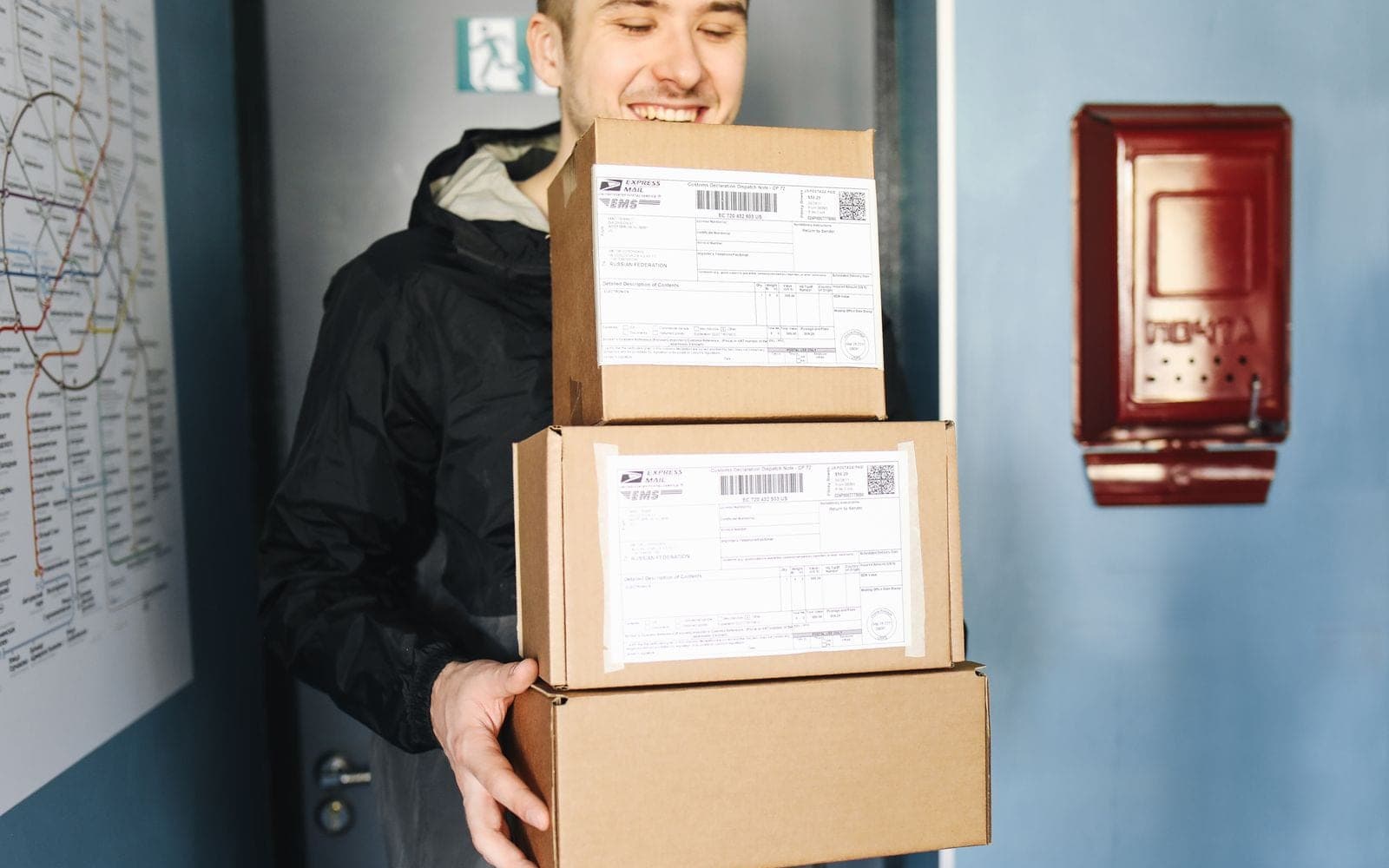 Entregador sorridente carrega três caixas de encomenda diante de uma porta