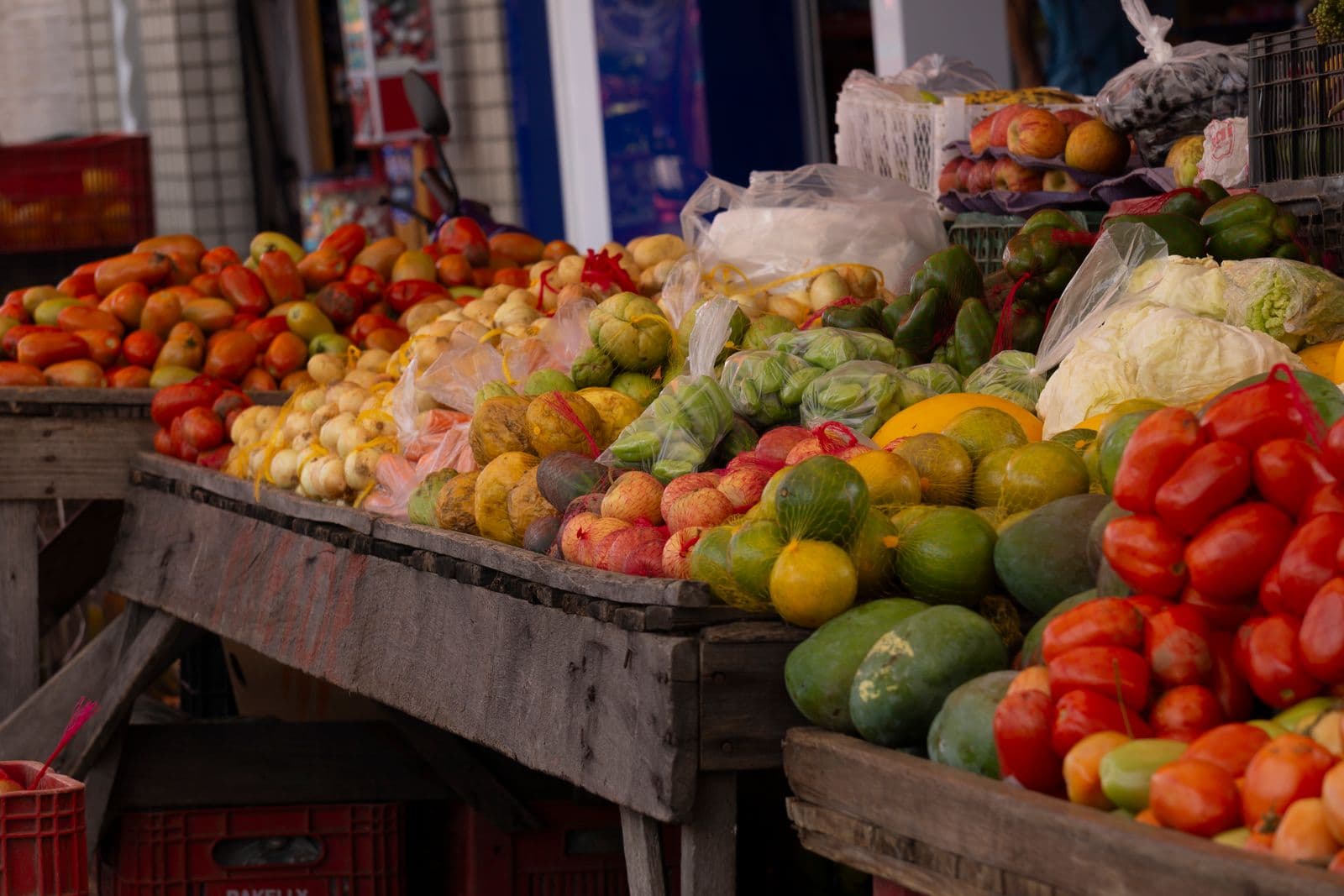 Banca de feira com tomates, cebolas, maracujás, abacates e pimentões em bancadas de madeira