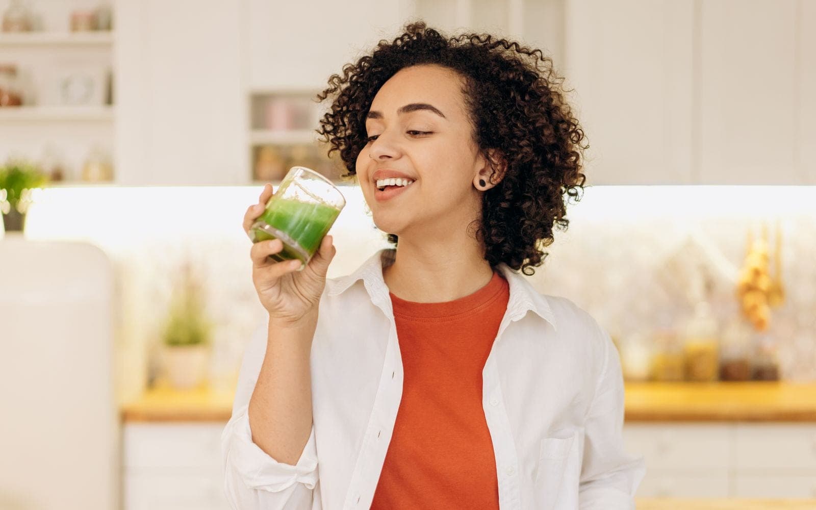 Mulher de cabelo cacheado sorri na cozinha segurando um copo de suco verde