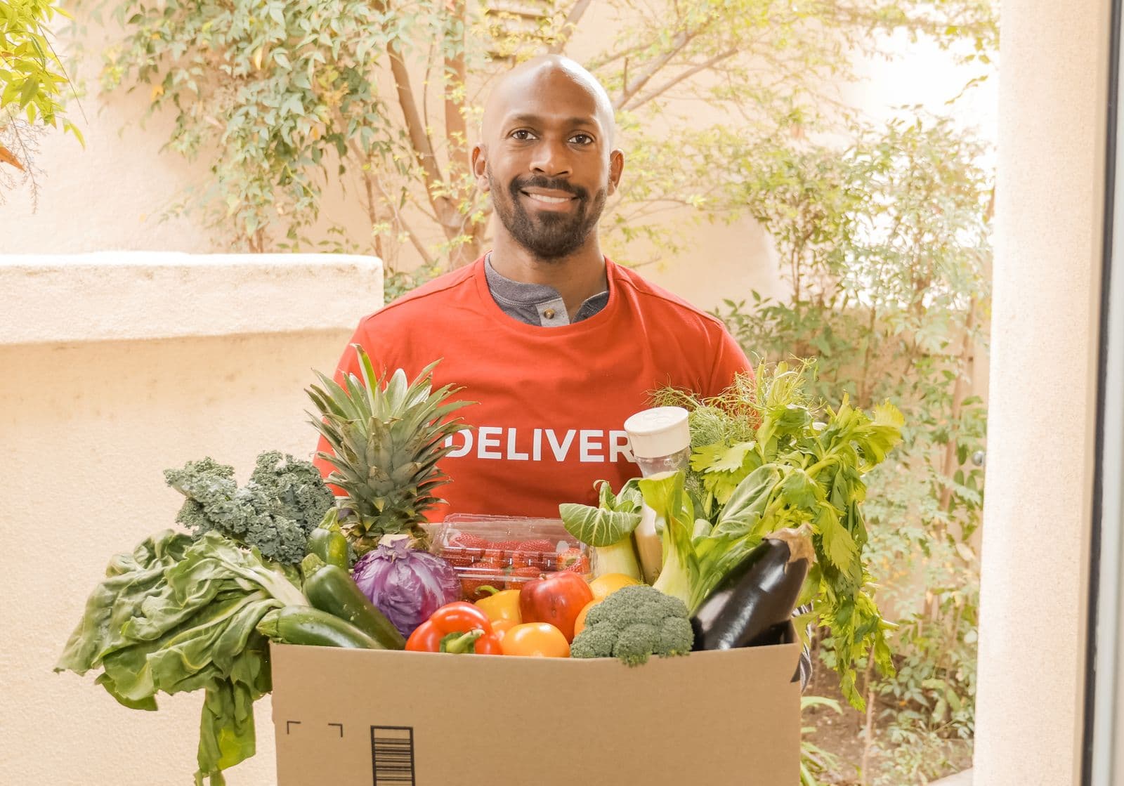 Entregador sorrindo entrega uma caixa de frutas e verduras frescas na porta de casa