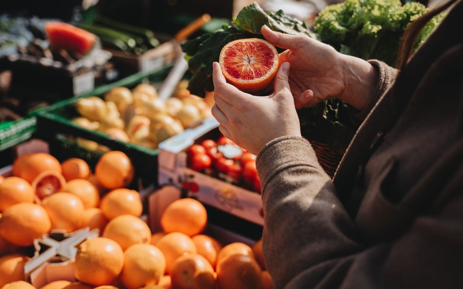 Mão segurando uma laranja sanguínea cortada diante de uma banca com laranjas e batatas