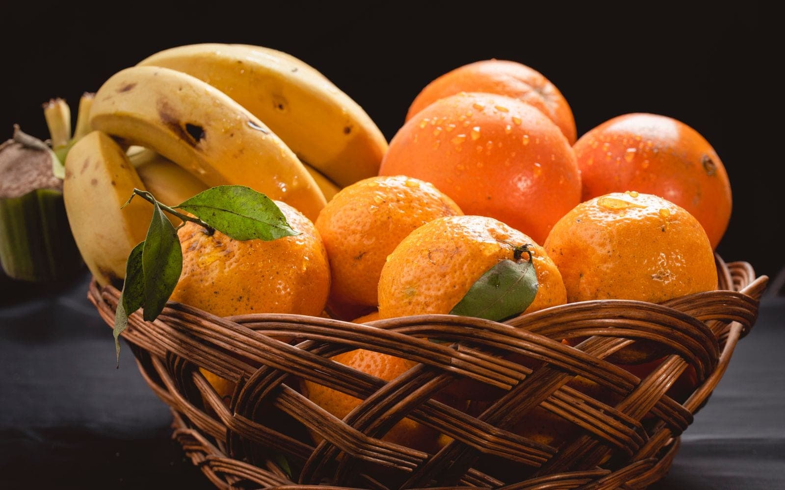 Cesta de vime com bananas, laranjas e tangerinas sobre uma mesa escura