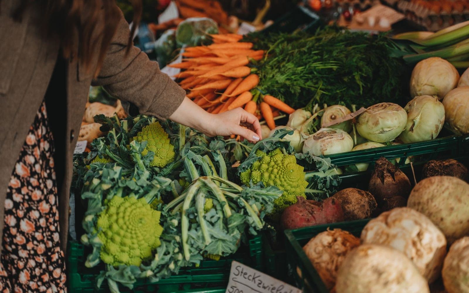 Mão escolhendo legumes numa banca, com cenouras, couve-flor romanesco e vagens à mostra