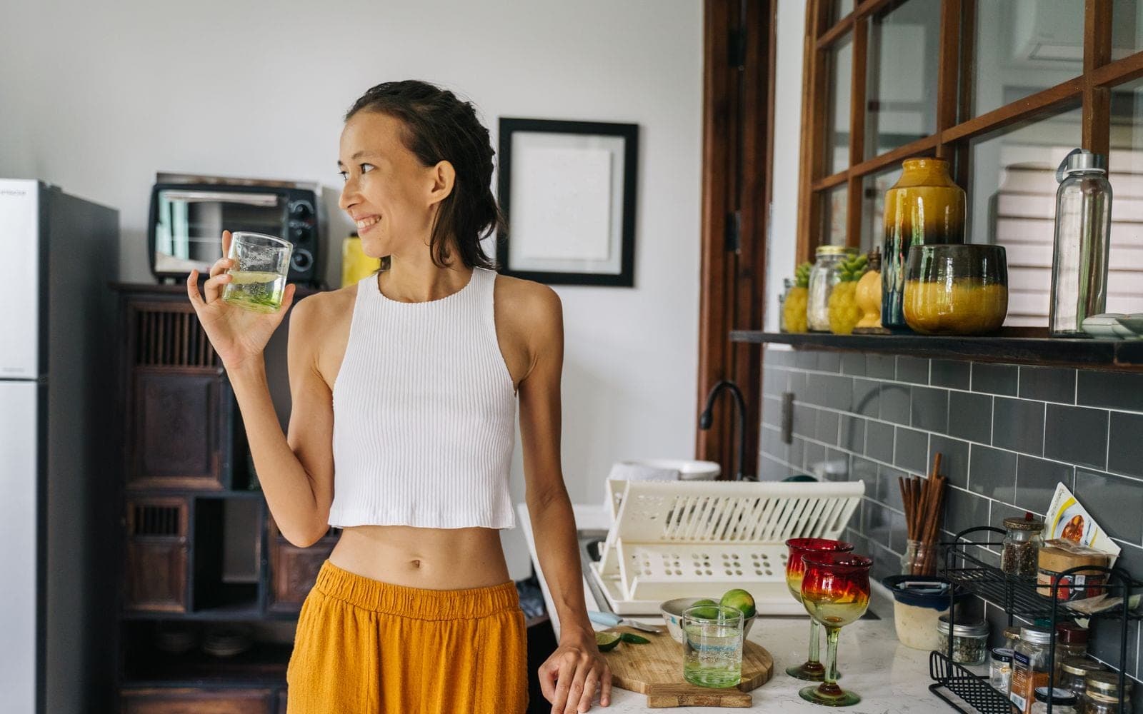 Mulher de top branco sorrindo ao segurar um copo numa cozinha com bancada e prateleiras