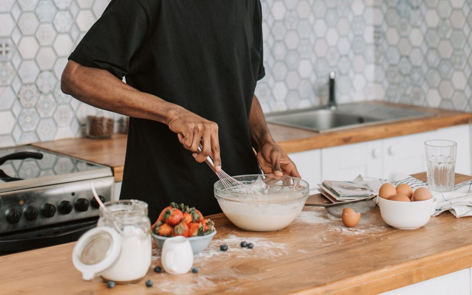 Homem de camiseta preta preparando comida numa cozinha clara com azulejos hexagonais
