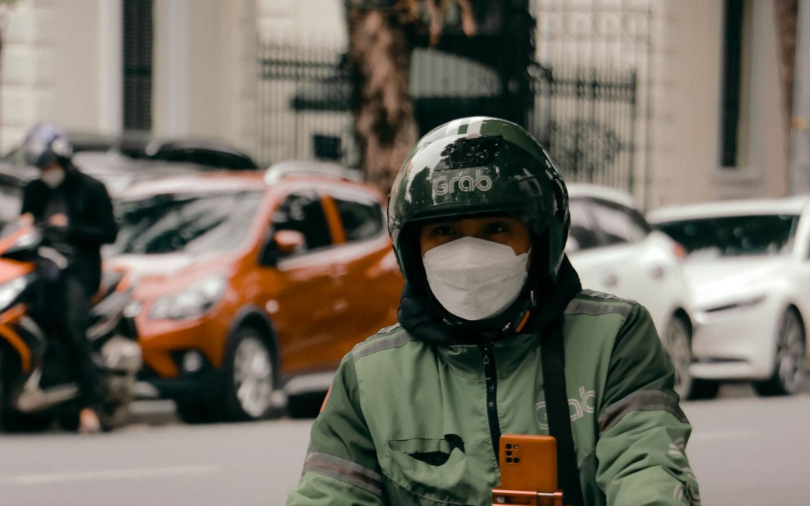 Entregador de jaqueta verde numa moto com celular no suporte e uma sacola de papel pendurada