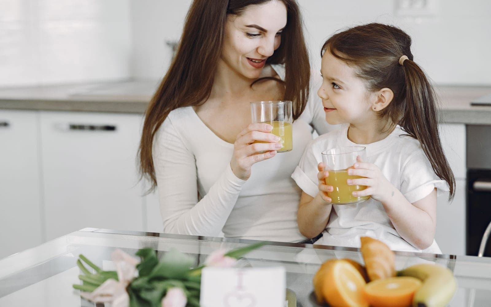Mãe e filha na cozinha brindam com copos de suco, com frutas ao fundo
