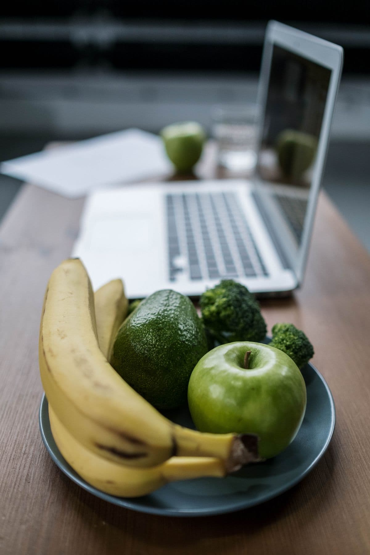 Prato com banana, abacate, maçã verde e brócolis ao lado de um notebook