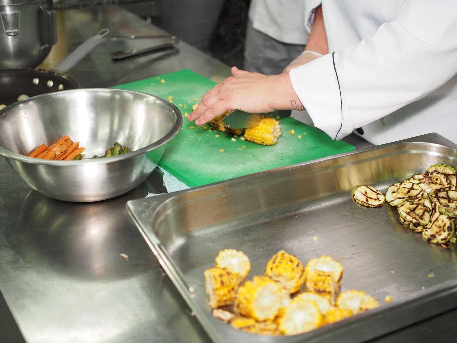 Cozinheiro cortando milho grelhado numa cozinha profissional, com abobrinhas grelhadas numa cuba