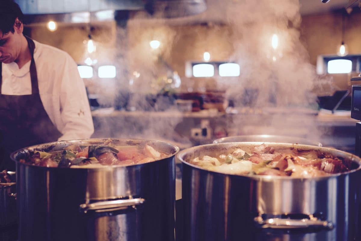 Cozinheiro ao lado de duas panelas grandes com legumes cozinhando em cozinha de restaurante