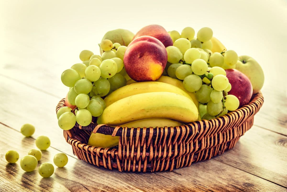 Cesta de vime com uvas, bananas e maçãs sobre mesa de madeira