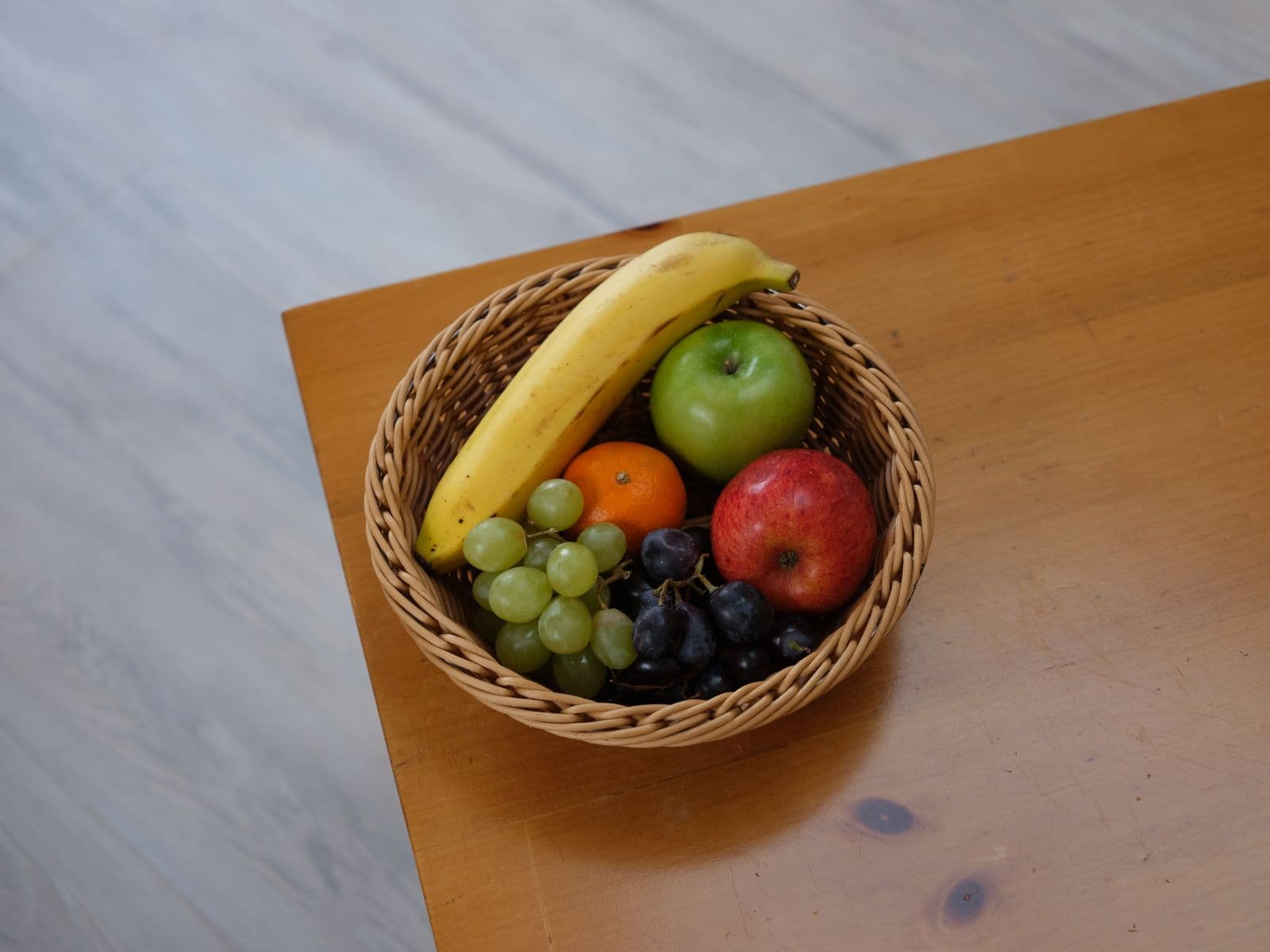 Cesta de vime com banana, maçãs, tangerina e uvas sobre mesa de madeira