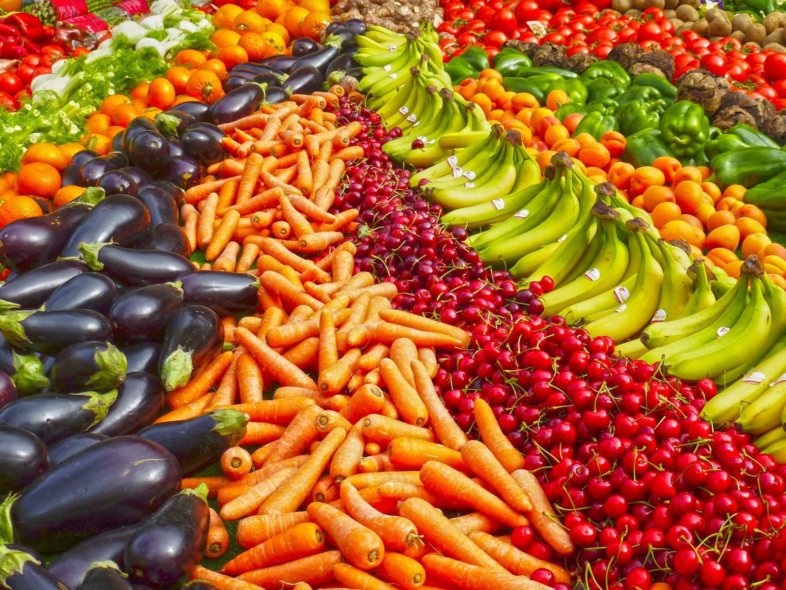 Fileiras coloridas de berinjelas, cenouras, cerejas, bananas e pimentões numa banca