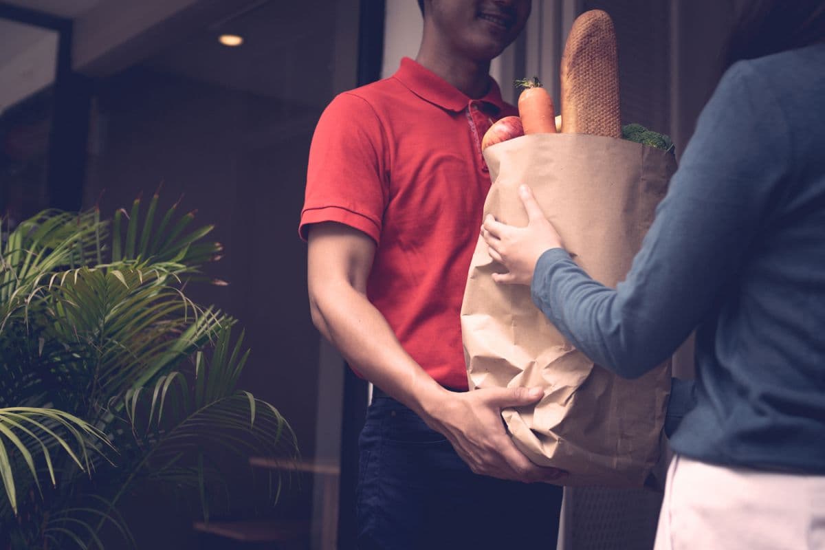Entregador de camisa vermelha entregando uma sacola de papel com frutas, verduras e pão