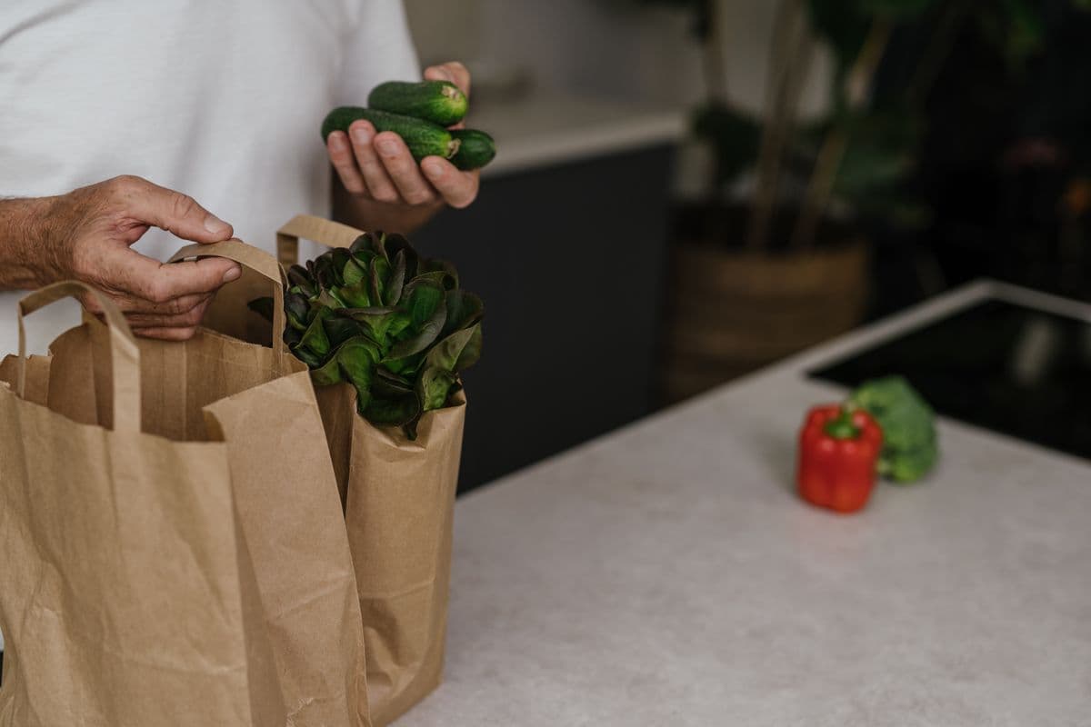 Mão tirando pepinos e alface de sacolas de papel sobre a bancada da cozinha