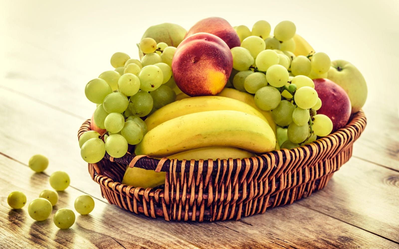 Cesta de vime com uvas, bananas, pêssegos e maçãs sobre uma mesa de madeira