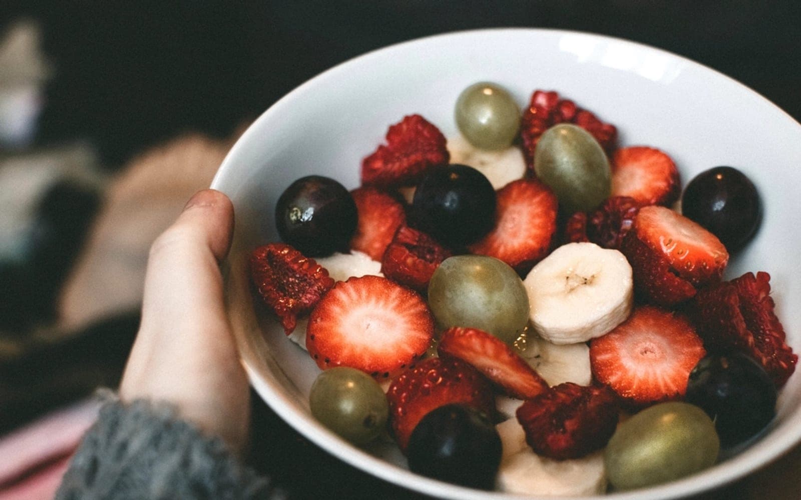 Mão segura uma tigela com morangos, uvas, banana e outras frutas picadas