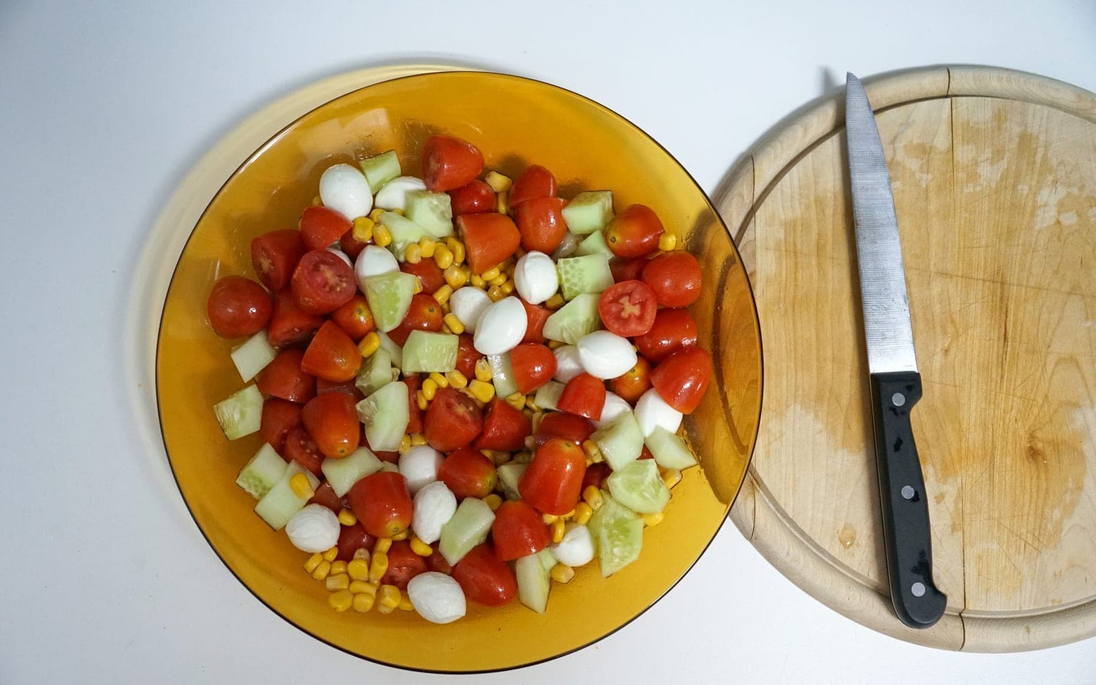 Tigela de vidro com salada de tomate, milho e pepino ao lado de uma tábua com faca