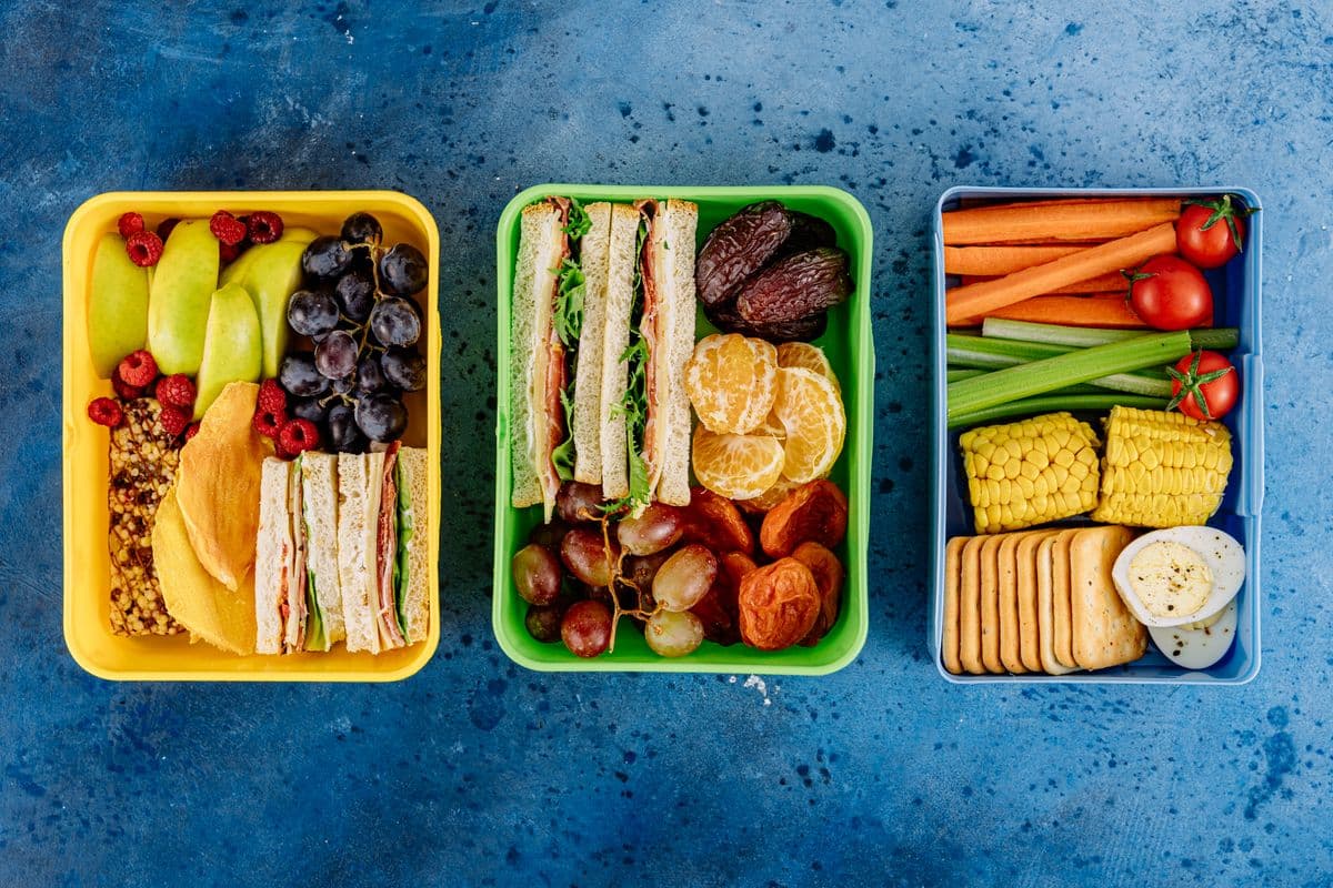 Três marmitas coloridas lado a lado, com frutas, sanduíches e vegetais organizados