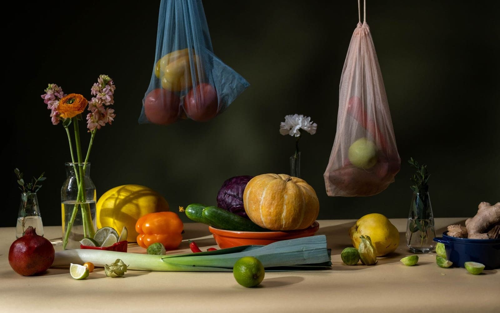 Mesa escura com abóboras, alho-poró, pimentões, flores e sacolas de tela com frutas penduradas