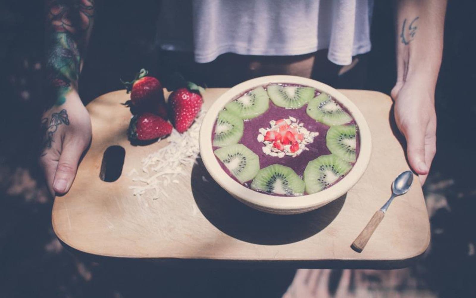 Duas mãos seguram uma tábua com uma tigela de frutas e fatias de kiwi