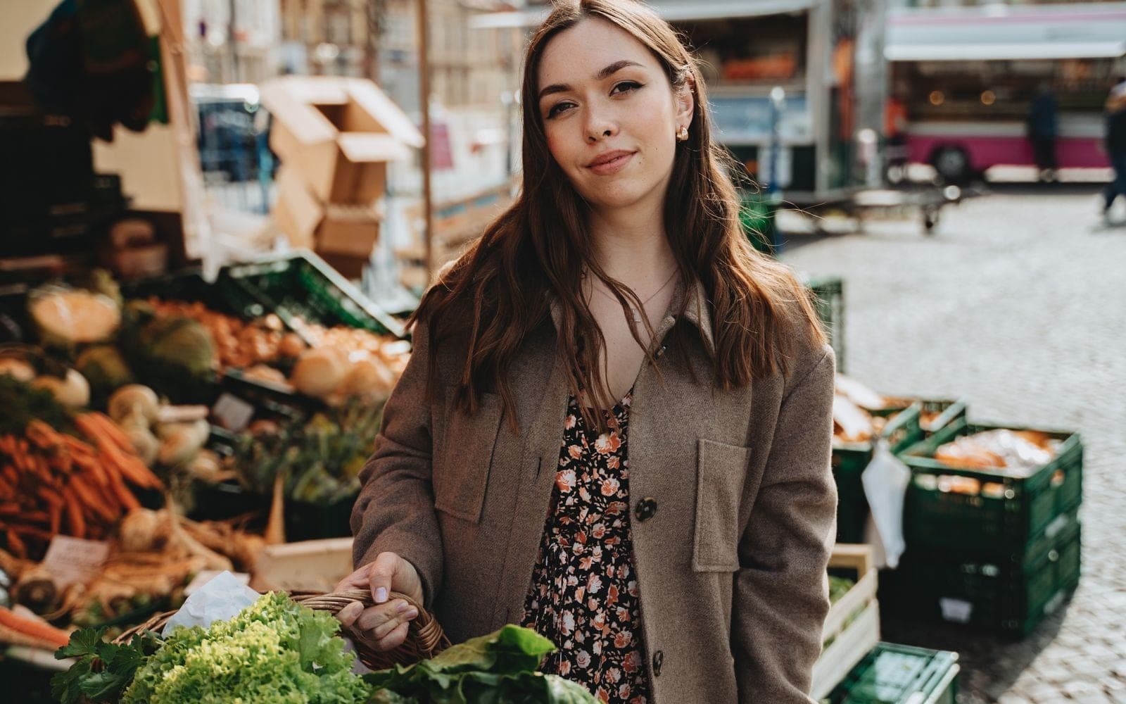 Mulher de casaco marrom posando ao lado de uma banca de verduras numa feira de rua