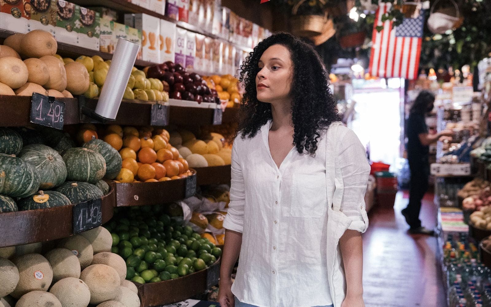 Mulher de camisa branca caminhando entre bancas de frutas e verduras em um mercado