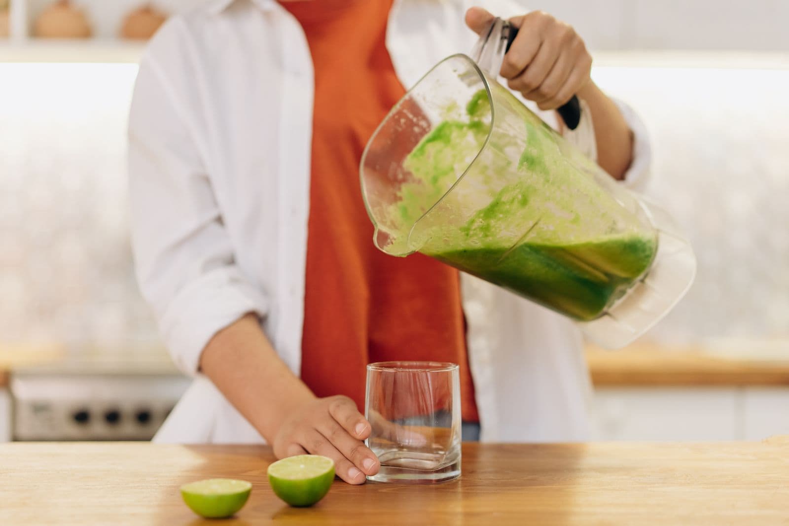 Pessoa servindo suco verde de um jarro de liquidificador em um copo, com limões cortados ao lado