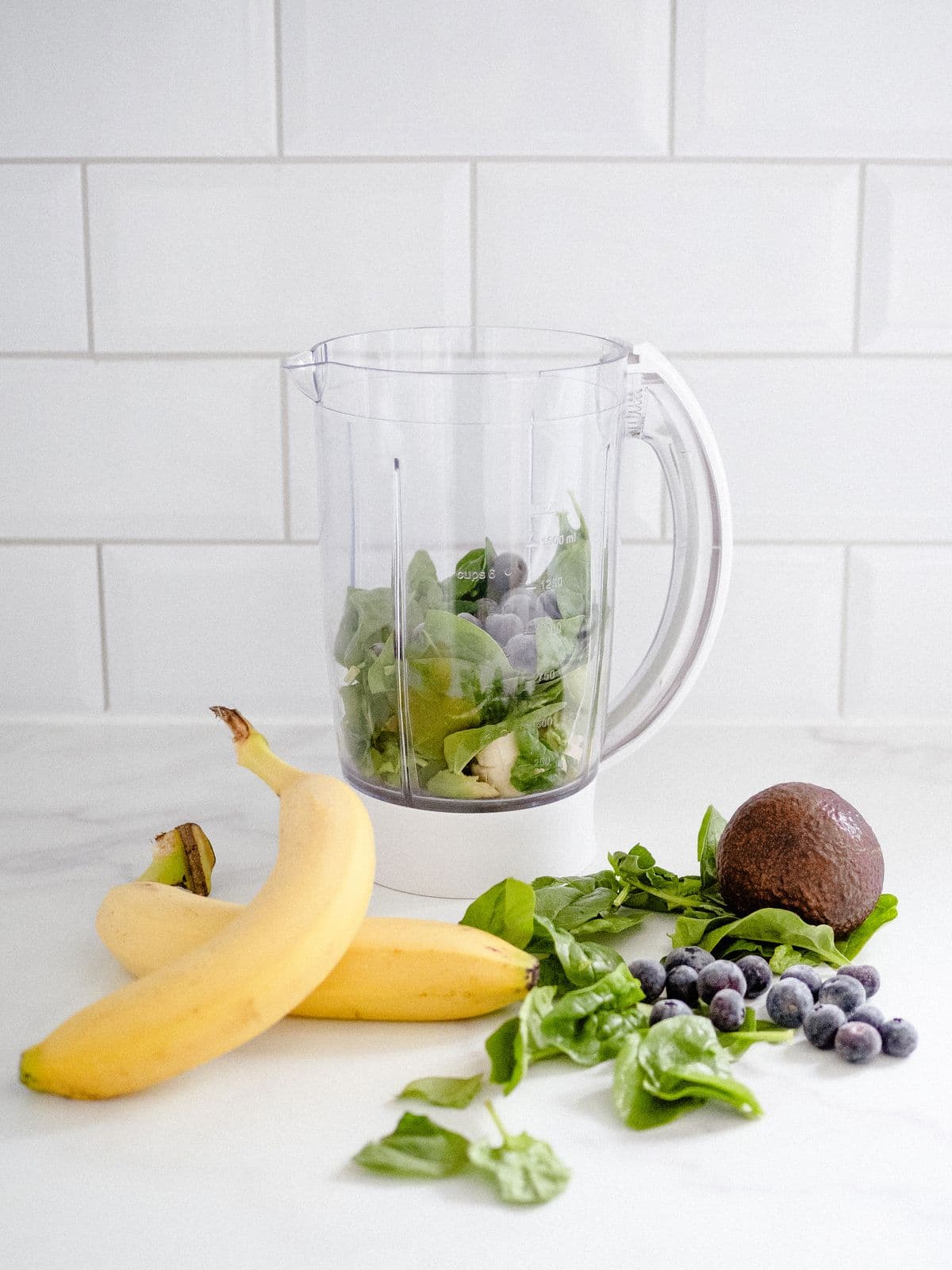 Liquidificador com folhas de espinafre, mirtilos e ingredientes prontos para bater, ao lado de bananas